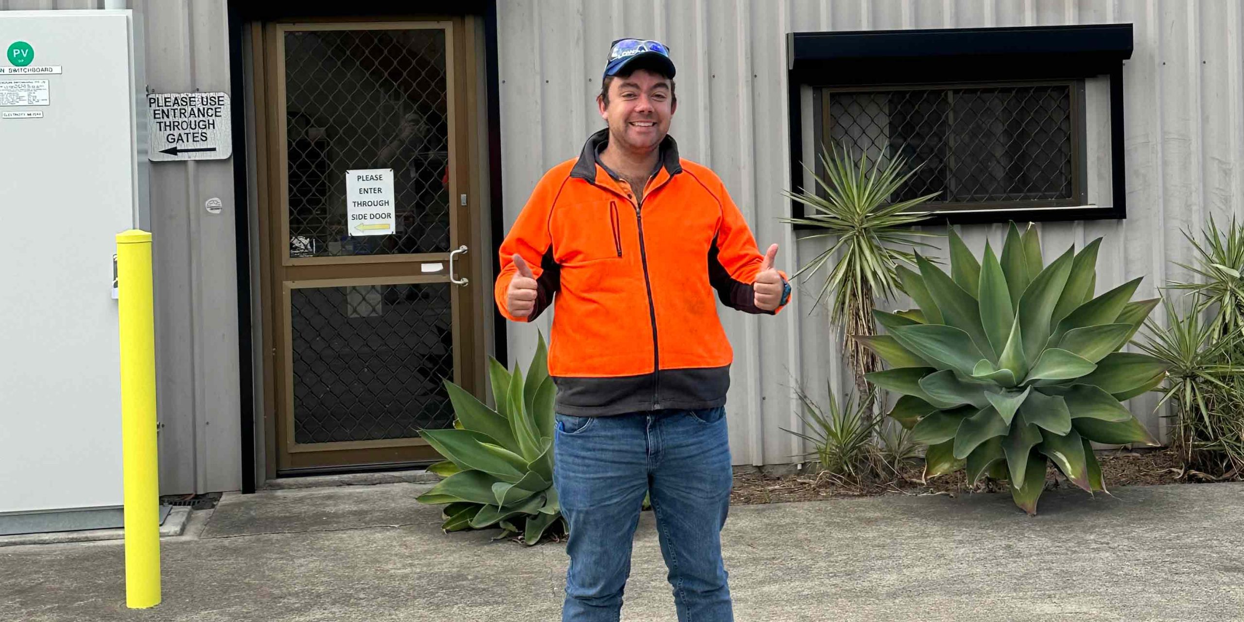 Man with two thumbs up and wearing hi-vis, standing in front of a shed.