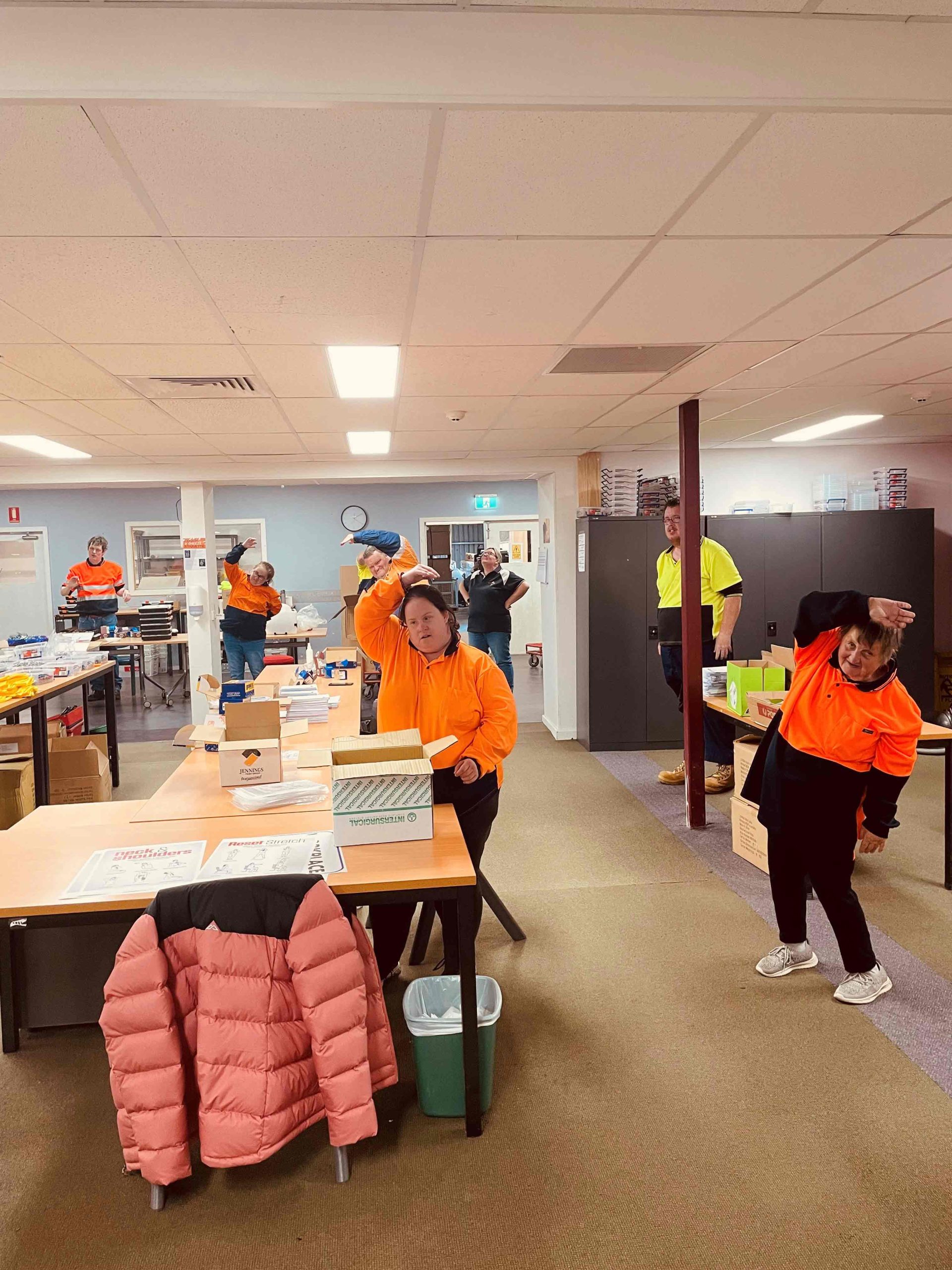 A room of men and women wearing high-vis stretching with one arm over the head.